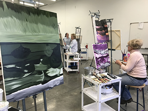 Photograph of a painting studio with multiple easels, work-in-progress paintings, and students.